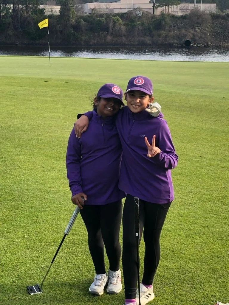 Two women in purple jackets and hats on a golf course.